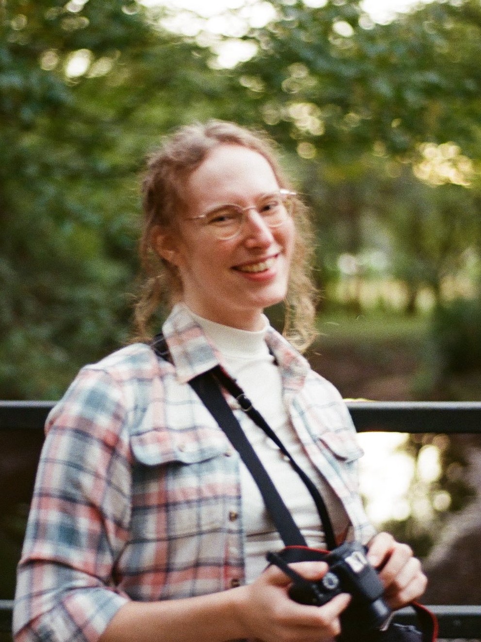 Allison Montgomery, a white woman, is looking at the camera and smiling. She is standing on a bridge over a creek wearing a light red, white, and blue flannel over a white tank top. She is holding her own camera. She looks very electable, and like she wants to preserve the natural environment.