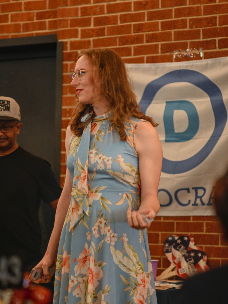 Allison Montgomery, a white woman, is speaking to an audience about policy issues. She has long, sandy brown hair and is wearing a light blue floral dress. Behind her is a Democratic party insignia. She looks especially electable in this one.