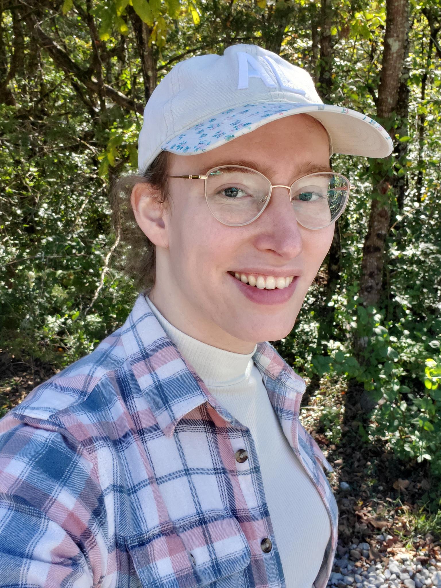 A photo of Allison Montgomery, a white woman, looking at the camera and smiling. She is in a wooded area, probably a hiking trail. She is wearing a white floral ball cap with an embossed "AL". light red, white, and blue flannel over a white tank top. She looks very electable.